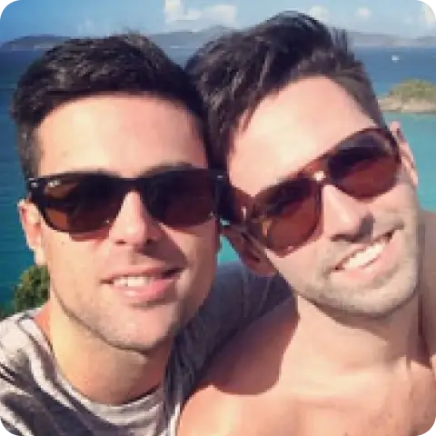 Billy and Pat smiling together in sunglasses, with a clear blue sky and ocean in the background.