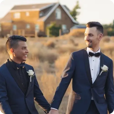 Bryan and Eddie holding hands and smiling at each other, dressed in elegant dark blue suits with boutonnieres, at an outdoor wedding setting.