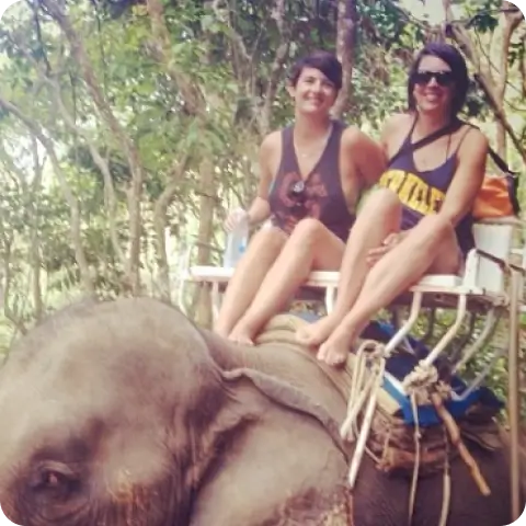 Maya and Kelly sitting on an elephant during an outdoor adventure, both smiling broadly.